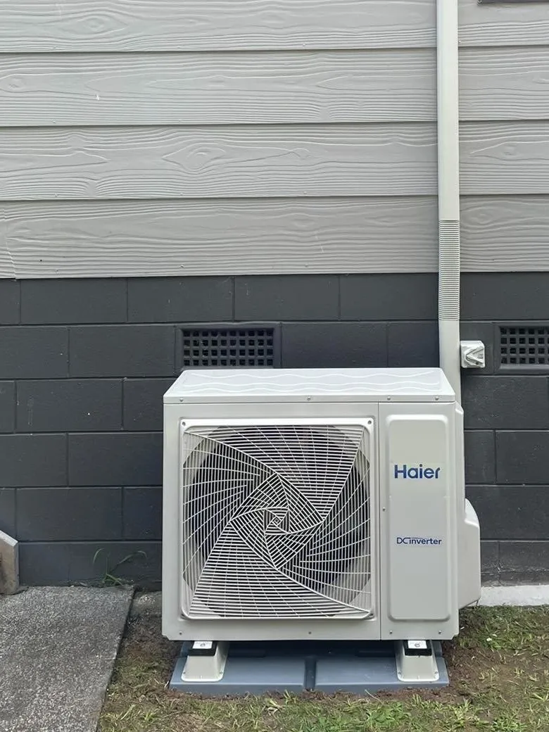 A heat pump outdoor unit on pads beside a grey weatherboard wall, with a white pipe cover running up the wall.
