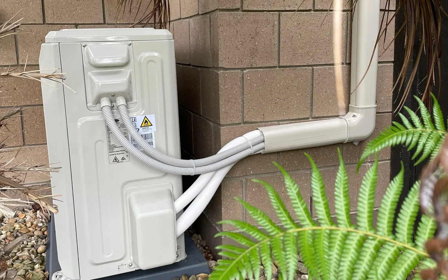 A heat pump outdoor unit against a block wall, with its pipes running into a white cover up the wall.