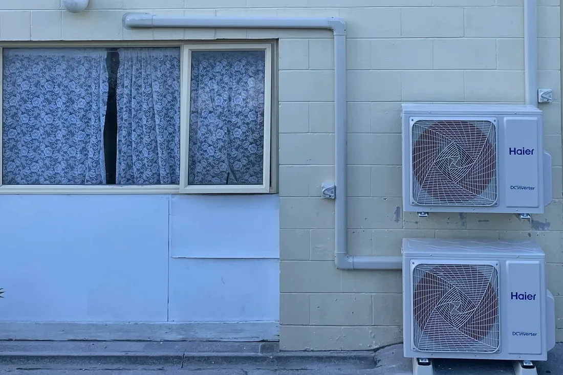 Two heat pump outdoor units, one above the other, beside a window on a painted block wall.
