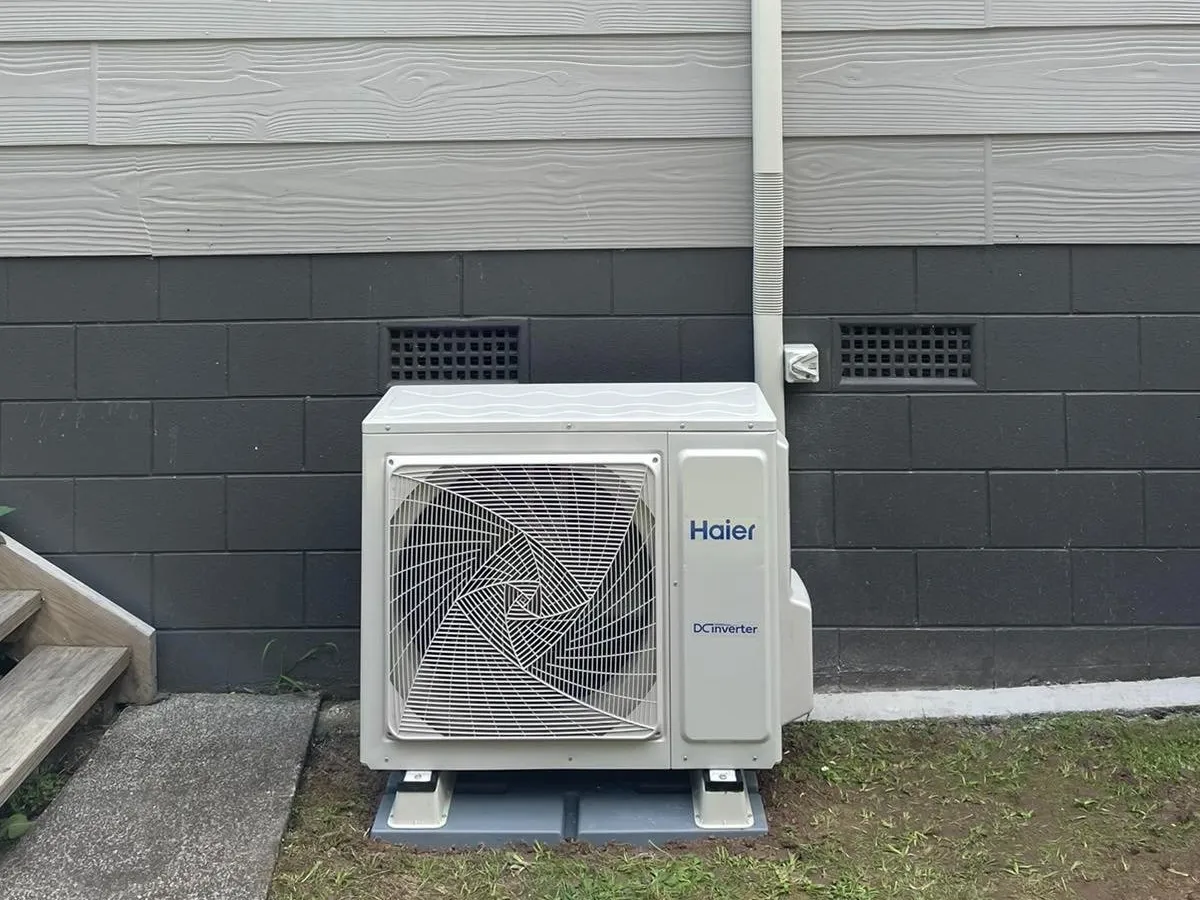 A heat pump outdoor unit on pads beside a grey weatherboard and block wall.